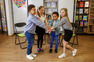 Four children stand in a circle inside a classroom, each with one hand stacked in the center as if starting a team cheer. They are smiling and appear to be engaged in a group activity. The room has bookshelves, folding chairs, and colorful artwork on the walls.