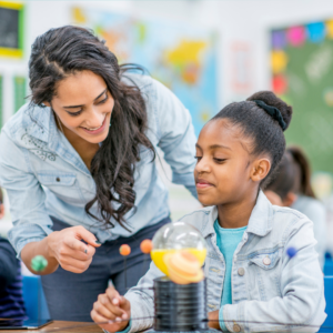 A smiling teacher works with a young student at a classroom table. The student is using a colorful hands-on model of the solar system while the teacher guides the activity.