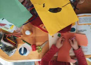 A child sits at a table working on a colorful craft project. Their hands are holding orange construction paper and a crayon. The table is scattered with supplies including scissors, glue, crayons, tape, and other pieces of colored paper. An open book with illustrations and text lies nearby.