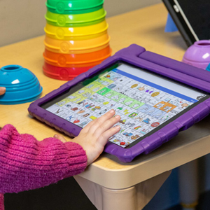 Close-up of a child's hand touching a tablet-based augmentative and alternative communication (AAC) device in a purple protective case. The screen displays a grid of picture-based communication symbols. Colorful stacking toys sit on the table nearby.