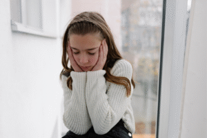 A young girl with long brown hair, wearing a white knit sweater and black pants, sits near a window with her eyes closed and her hands pressed to the sides of her face. She appears deep in thought or distressed. The setting is bright with natural light coming through the window.