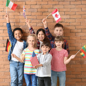 Six smiling children stand together in front of a brick wall, holding flags representing Italy, the United Kingdom, Canada, Germany, the United States, and Lithuania.