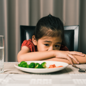 A young child sits at a dining table with their head resting on folded arms, looking down at a plate of vegetables that includes broccoli and carrots. A glass of water sits nearby, and the child appears reluctant or uninterested in eating the food in front of them.