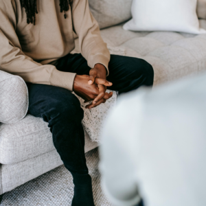 A close-up photo of an adult seated on a light-colored couch with their hands clasped in their lap during a therapy session. The therapist is visible out of focus in the foreground, creating the perspective of a conversation in a calm, comfortable office setting.