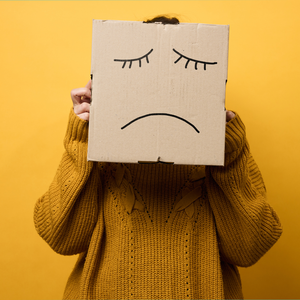 Person wearing a mustard yellow sweater holds a cardboard box in front of their face. A simple sad face with closed eyes and a frown is drawn on the box against a bright yellow background.