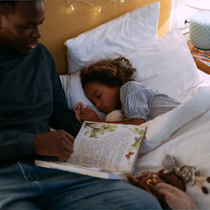 A parent sits beside a sleeping child tucked into bed while holding an open storybook. The child rests against a pillow with a stuffed animal nearby.