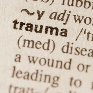Close-up photograph of the word “trauma” printed in bold black letters in a dictionary, with portions of its pronunciation, definition, and surrounding text visible.